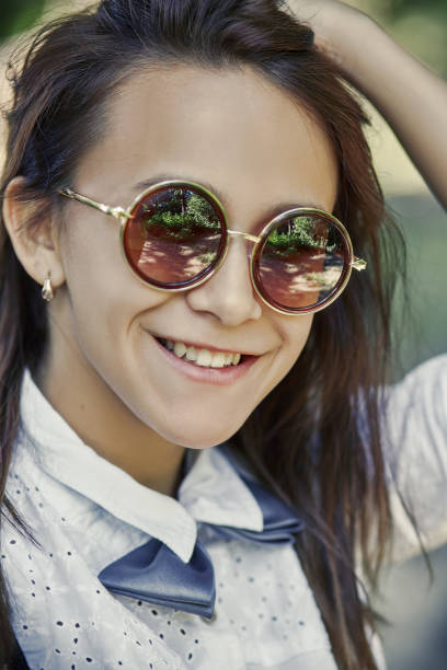 Young woman in strapped top wearing sunglasses and striped bandana looking at camera with cheerful smile, enjoying cool weather and sun whilst on vacation with blurry Tuscan landscape lined with cypress trees in background