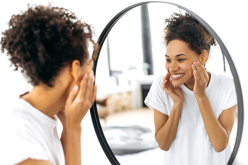 Joyful African American curly girl in white t-shirt, stands in front of a mirror in the living room, smiles happily looking at her face in the mirror, is pleased with the condition of her facial skin