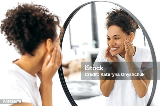 istock Joyful African American curly girl in white t-shirt, stands in front of a mirror in the living room, smiles happily looking at her face in the mirror, is pleased with the condition of her facial skin 1415985075