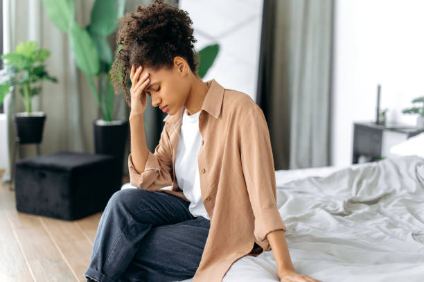 Upset african american girl, stylishly dressed, sit on bed at home, preoccupied with personal problems, sad by bad news, experiencing depression, despair, headache,needs rest and psychological support stock photo