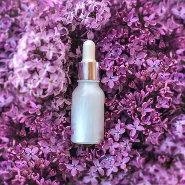 White frozen glass cosmetics dropper bottle on lilac flowers in blossom. Skin care, natural beauty products presentation concept. Romantic composition. Flat lay, top view