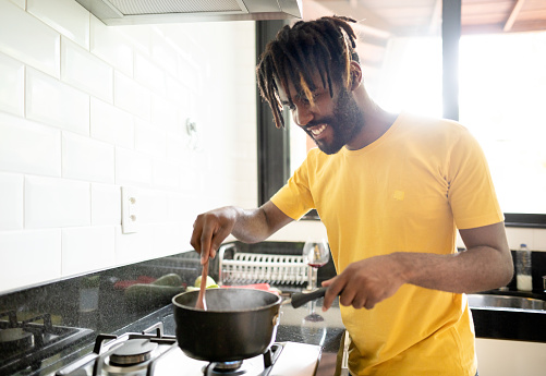 Smiling young man stirring sauce in a cooking pot while preparing dinner on his kitchen stove at home