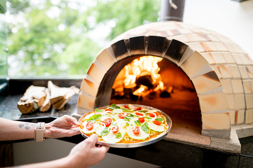 Close-up of a woman putting a freshly-made pizza into an oven outside on a patio