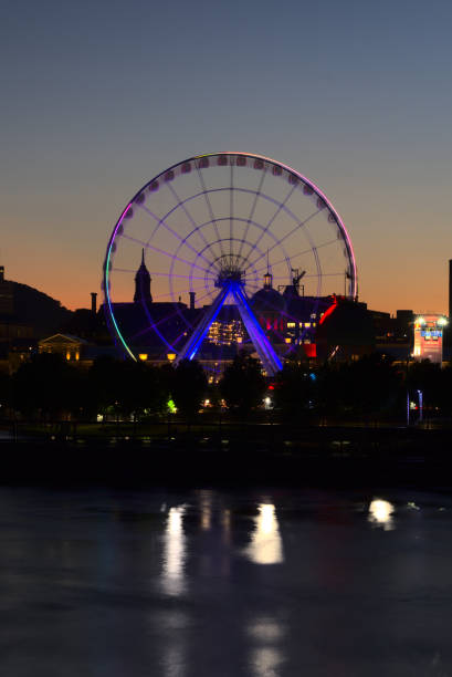 Long exposure of Montreal Grand Ferris Wheel in Old Port with motion blurat twilight blue hour. Reflections of sun rays on Saint Laurent river with a background of cloudy blue sky stock photo