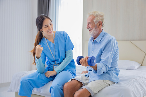 Hospice nurse is helping Caucasian man in bed to exercising muscle strength in pension retirement center for home care rehabilitation and post recovery process