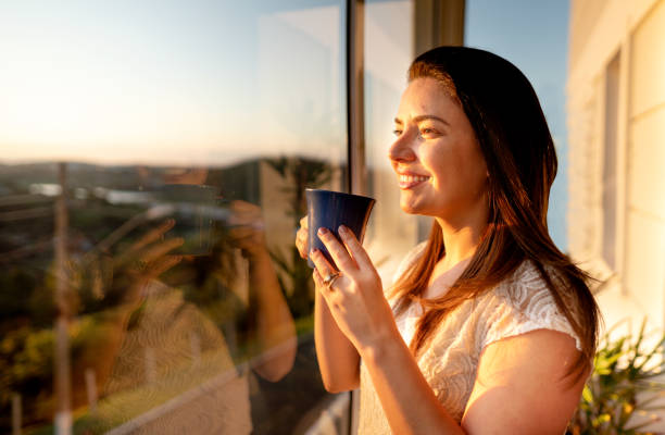 Young woman drinking coffee and watching the sunset through a window at home