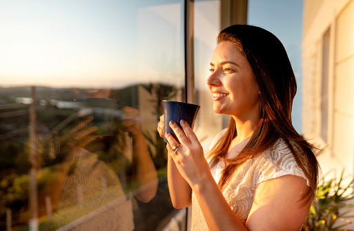 Young woman drinking a cup of coffee while looking out at the sunset through a window at home