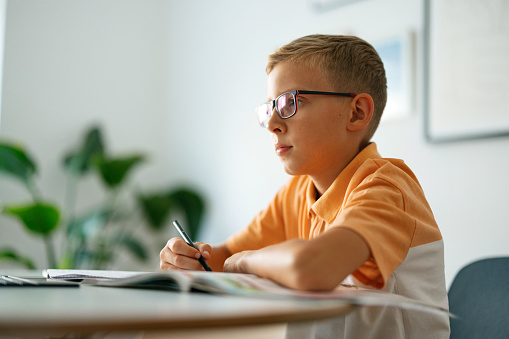 Cute young blonde boy sitting at the table, wearing glasses and studying. He is looking in front of him. There is an open book on the table. He is looking serious and focused.
