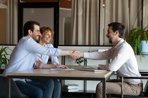 Happy millennial married couple shaking hands with hispanic real estate agent, feeling satisfied with professional service, making deal celebrating signing paper contract agreement at office meeting.