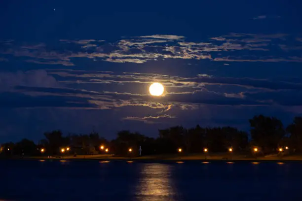 Supermoon as seen from Lachine, Montreal, Quebec golden full moon rising over the water casting moonlight reflection on the water with illuminated clouds and star in sky on beautiful summer night sturgeon super moon photography