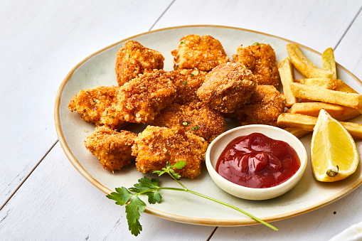 Chicken nuggets on a plate, with ketchup, french fries and lemon, served on a white wooden restaurant or home table