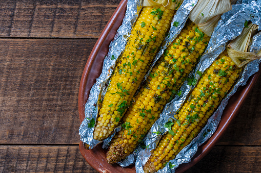 Tasty corn baked in foil with butter, garlic, chili, parsley and sea salt. Grilled corn in aluminum foil on brown wooden table. Top view, close up, copy space. Healthy food concept