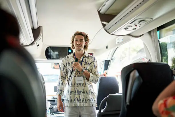 Waist-up view of smiling man in relaxed attire standing at front of bus and discussing travel details with seated passengers.