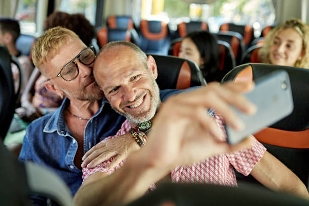 Candid portrait of smiling gay couple taking selfie on coach Head and shoulders view of bearded vacationers in 40s and 50s embracing and looking at smart phone as they capture a travel memory. tourist couple candid travel stock pictures, royalty-free photos & images