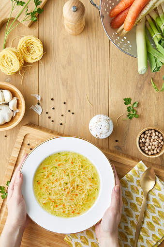 The woman hands holding a vegetable soup plate above the kitchen, wooden table, with fresh vegetables, ingredients and seasoning around the center, top view with a free space for copy in the middle