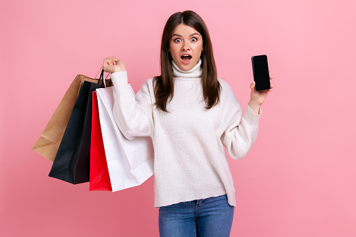 Astonished dark haired female posing with shopping bags, showing phone with blank screen for ad, wearing white casual style sweater. Indoor studio shot isolated on pink background.