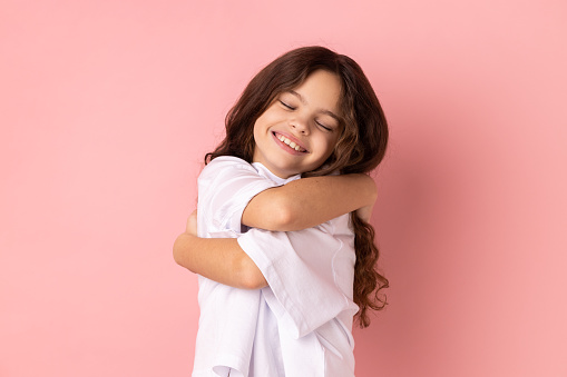 Portrait of satisfied happy little girl wearing white T-shirt embracing herself, standing with eyes closed from pleasure and happiness. Indoor studio shot isolated on pink background.