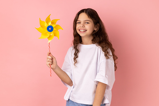 Portrait of satisfied delighted charming little girl wearing white T-shirt holding paper windmill, playing with pinwheel toy on stick. Indoor studio shot isolated on pink background.