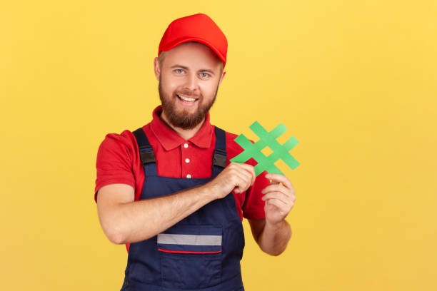 istock Workman wearing overalls standing, holding green hashtag, looking at camera with happy expression. 1416037892