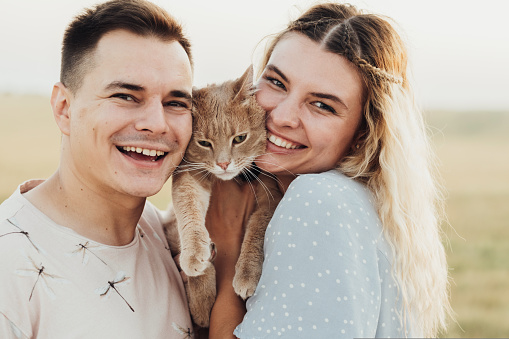Close Up Portrait of Cheerful Caucasian Couple with Their Cat, Young Woman and Man Hugging with Pet Outdoors at Sunny Summer Day