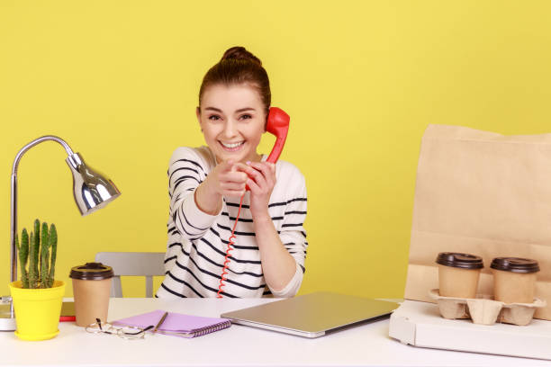 istock Woman talking on landline phone, pointing at camera, communicating with client sitting at workplace. 1416038135