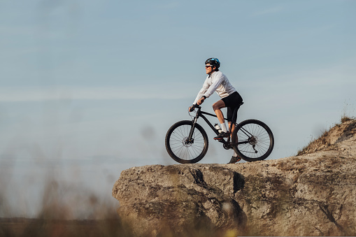 Equipped Professional Male Cyclist Standing with Mountain Bike on Edge of Rock, Sky on the Background and Copy Space