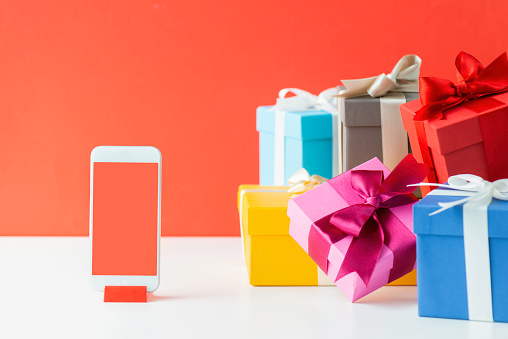 Multicolored gift boxes and a smart phone on white table in front of a red wall.