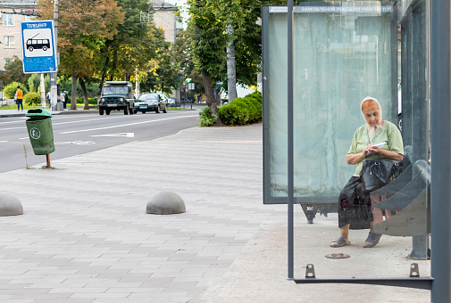Elderly woman sitting on a bench in pavilion of public transport stop. Ukraine, Zhytomyr, August 19, 2022