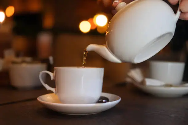White cup with teapot. The hand holding the teapot pours tea into a pot standing on saucer in soft focus on naturally blurred background. Coffee, tea house, bokeh lights. The concept of a cozy pastime