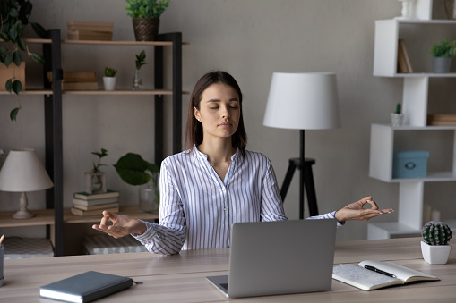 Relaxed peaceful young business woman sitting at workplace with folded hands in mudra gesture, meditating or breathing fresh air, relieving stress or enjoying peaceful break time in modern office.