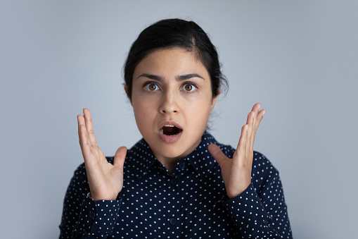Stressed surprised terrified young Indian ethnicity woman looking at camera with opened mouth, feeling confused getting unexpected news, isolated on grey studio background, fearful face expression.