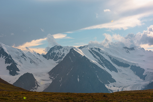 Scenic landscape with high snowy mountain range and long glacier tongue in sunset golden sunlight under cloudy sky. Awesome view to sunlit large snow mountains under sunset sky at changeable weather.