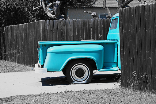 The passenger side view of the bed of a vintage pickup truck.   Light blue pickup truck.  Flat tire.   Photograph with blue filter - pickup truck in color and the rest of the photograph in black and white.