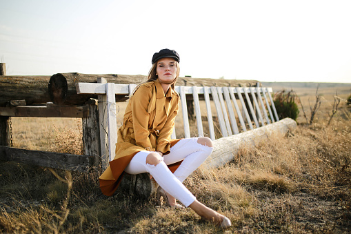 Beautiful young blonde woman sitting on the floor in an orange trench coat taken at the horse farm . fashion shoot
