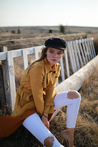 Beautiful young blonde woman sitting on the floor in an orange trench coat taken at the horse farm . fashion shoot