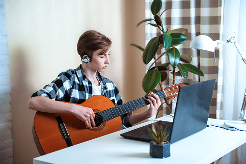 Teenage boy is learning to play guitar by taking lessons using laptop. Additional online education