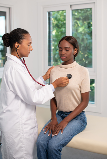 Focused African American woman at appointment. Female patient sitting on sofa, doctor using stethoscope. Medicine, health concept