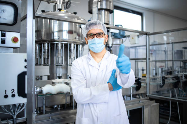 portrait d’un technologue ou d’un travailleur vêtu de vêtements blancs stériles debout près d’une machine industrielle automatisée dans une société pharmaceutique ou une usine. - équipement de production pharmaceutique photos et images de collection