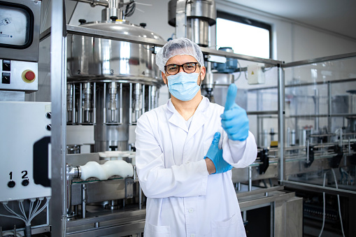 Portrait of technologist or worker in sterile white clothing standing by automated industrial machine in pharmaceutical company or factory.