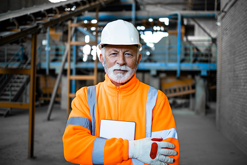 Portrait of an experienced senior industrial worker in safety equipment standing in factory interior.