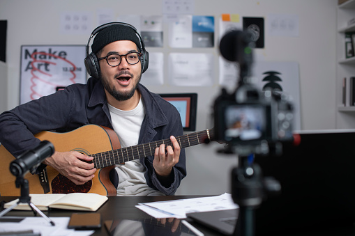 Young Asian male graphic designer blogger influencer playing guitar talking looking at camera shooting education tutorial vlog training filming video course for social media at studio.