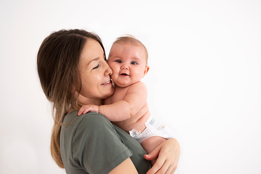 A smiling mom is standing and holding a beautiful baby in her arms looking at the camera, behind them is a white background with plenty of copy space.