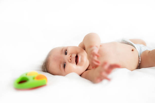 A beautiful smiling and happy baby in diapers lies on her back on a clean white bed and looks and points with her hands to a toy in front of her.
