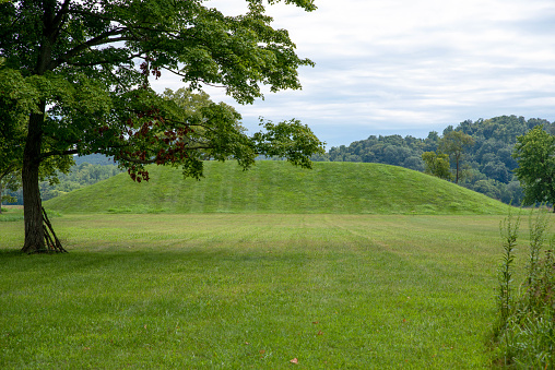 Grass covered prehistoric native American Hopewell culture burial mound at Seip Earthworks in Ohio under a dramatic cloudy sky with neatly cut green grass foreground and tree with green foliage and sticks leaning against the trunk. Woodland background. No people, with copy space.