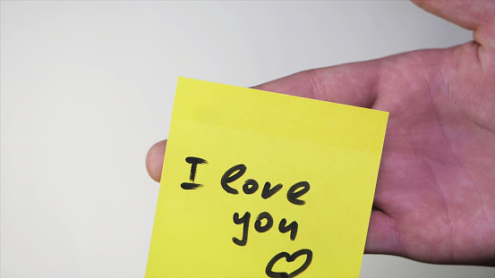 Note with I love you. Blank yellow sticky note with I love you sign pined on a cork bulletin board. Inscription on the sticker I love you on white background. Man's hand put blank yellow sticker isolated on a white background