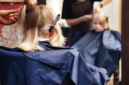 Hair salon, barber woman make fashionable pretty hairstyle for cute young blond child in modern barbershop. Hairdresser makes hairdo for little girl in barber shop. Concept hairstyle and beauty