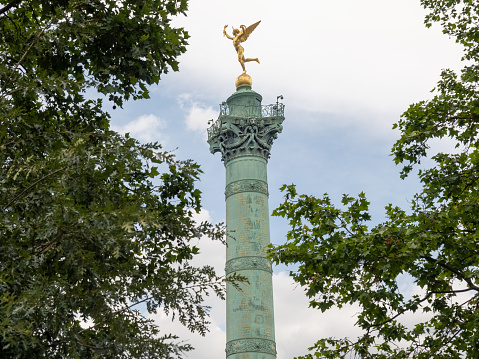 View of the July Column and the gilded figure of the Le Génie de la Liberté (Spirit of Freedom) on the top. In the photo, the memorial is framed by tree foliage on the right and left. 06/04/2022 - Pl. de la Bastille, 75004 Paris, 11th arrondissement, France