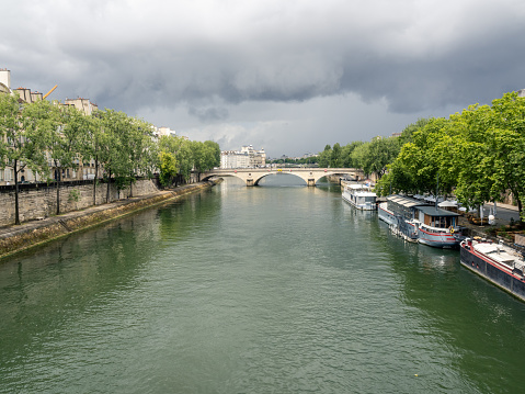 view of the old stone arch bridge over the river Seine, the bridge leads from the right bank of the Seine to the Île Saint-Louis. trees with green leaves on the right and left of the riverbank, under a cloudy sky. 06/08/2022 - Voie Georges Pompidou, 4th arrondissement, 75004 Paris, France
