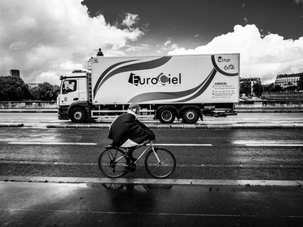 Black and white photo, in the foreground a cyclist with a rain cape driving to the left, in the background a truck with the inscription EUROCIEL driving to the right. The road is wet from the rain, the sky partly cloudy. 06/08/2022 - Pont d'Austerlitz, 75012 Paris, France