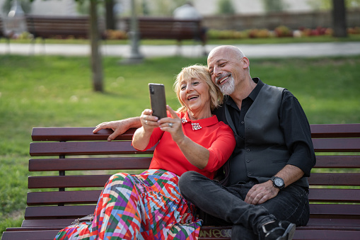 Man and woman, happy mature couple sitting on bench in public park outdoors in city, they are taking a selfie with smart phone.